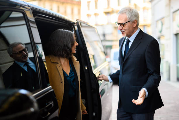 Senior chauffeur welcoming a guest beside a black executive vehicle outside a hotel